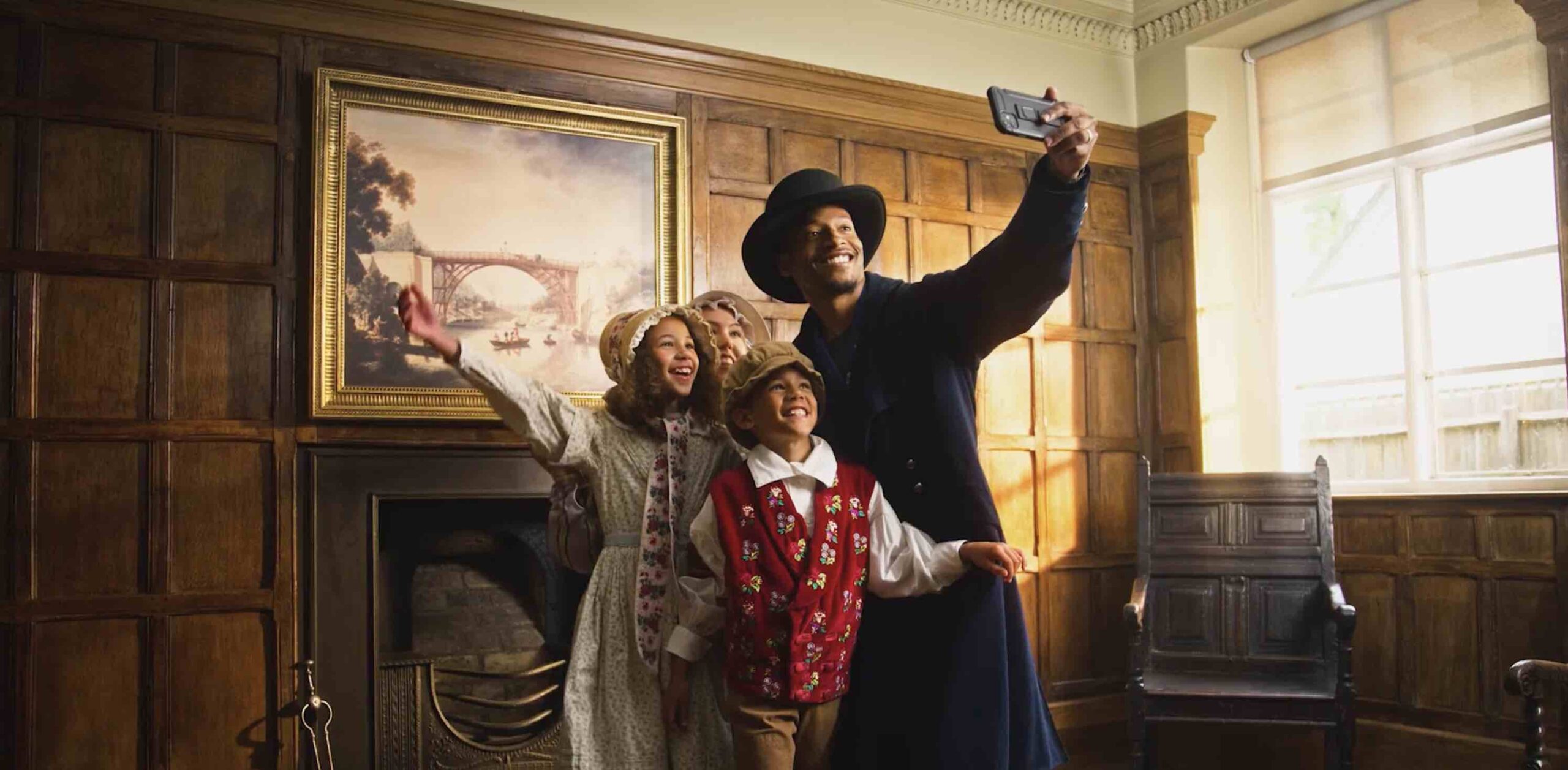 Family of 4 in Victorian clothes taking a selfie in front of painting of Ironbridge