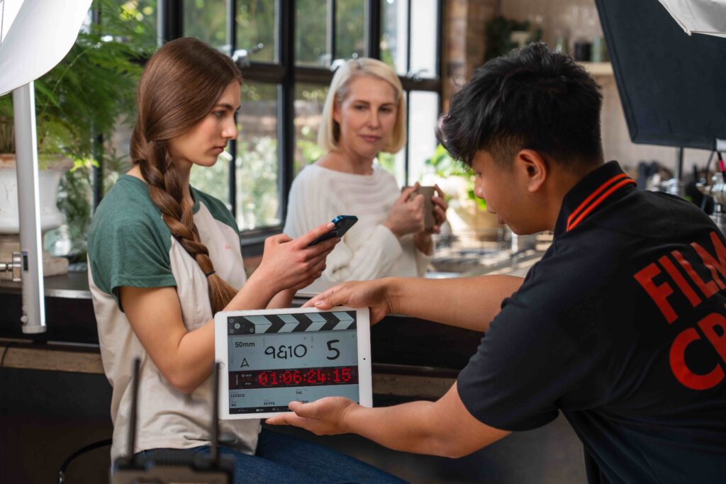 Camera assistant holding Clapperboard in front of Girl with plait wearing white and green t-shirt on her phone next to blonde woman holding mug. Shot for Luceco TV ad.