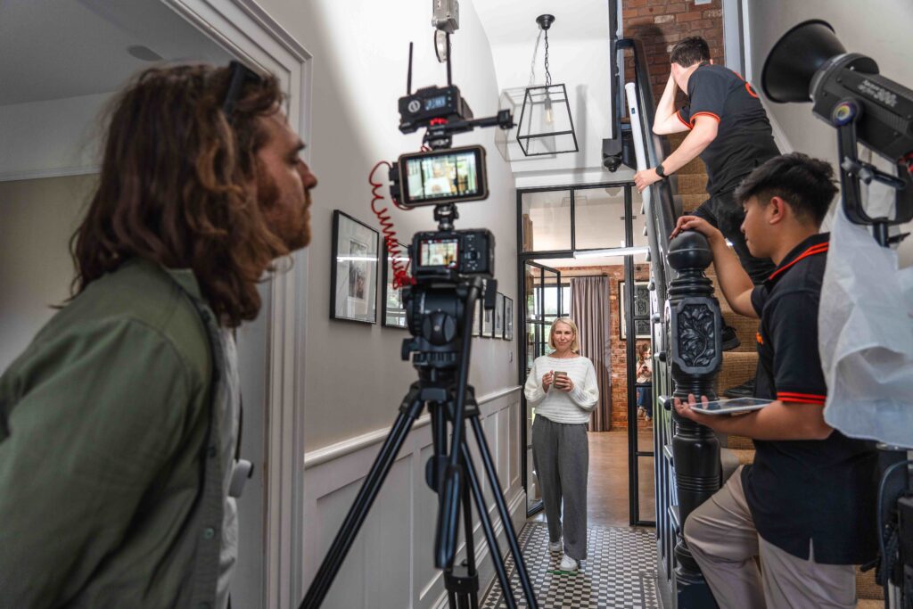 DOP looking at camera on tripod filming a blonde woman in a white jumper holding a mug walking down a hallway for Luceco TV ad