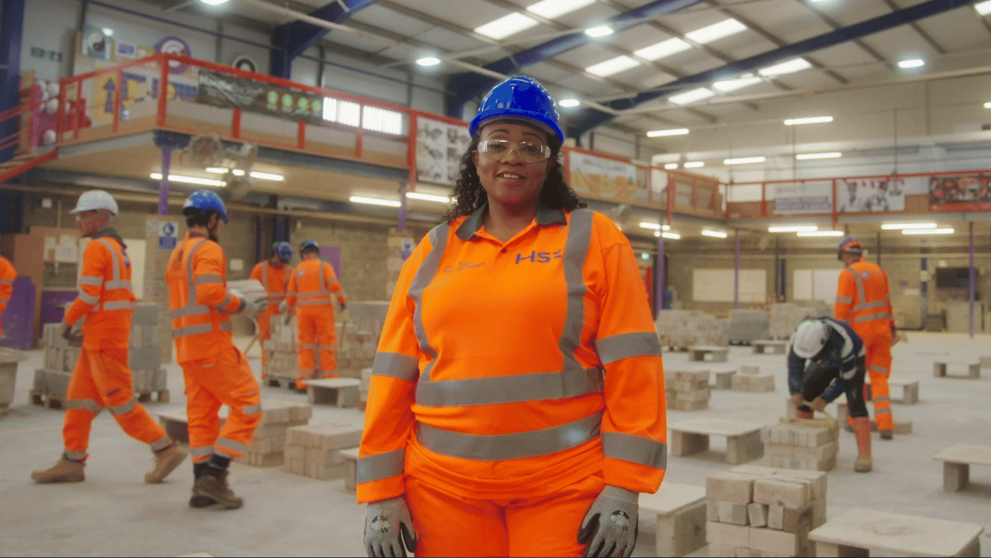 Woman who is in a warehouse facility wearing Hi-Vis. The South & City College Birmingham logo appears and transitions the scene into her wearing a helmet that says HS2 with the background being a HS2 construction site.