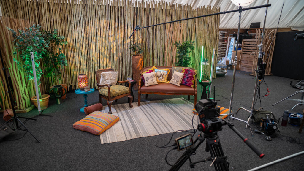 The set we built at West Midlands Safari park, featuring a bamboo wall, chairs, cushions, carpet and plants. Camera on a tripod filming the set up. Filming lights in the background.