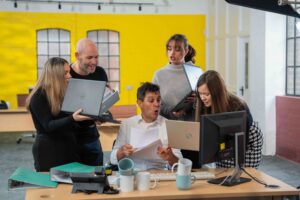 Behind the scenes at Technical Drive shoot. 1 office worker sat at desk, 4 others gathered around him holding their laptops looking stressed. Mugs piled up on the desk. Pure chaos. The background is an office environment with a yellow wall.