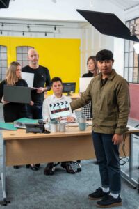 Camera Assistant holding clapperboard in front of 4 actors gathered around an office desk with a yellow background.