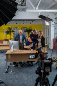 Behind the scenes at Technical Drive shoot. 1 office worker sat at desk, 4 others gathered around him looking happy. Background is vibrant office setting with yellow wall. Camera and lights set up to film the scene.