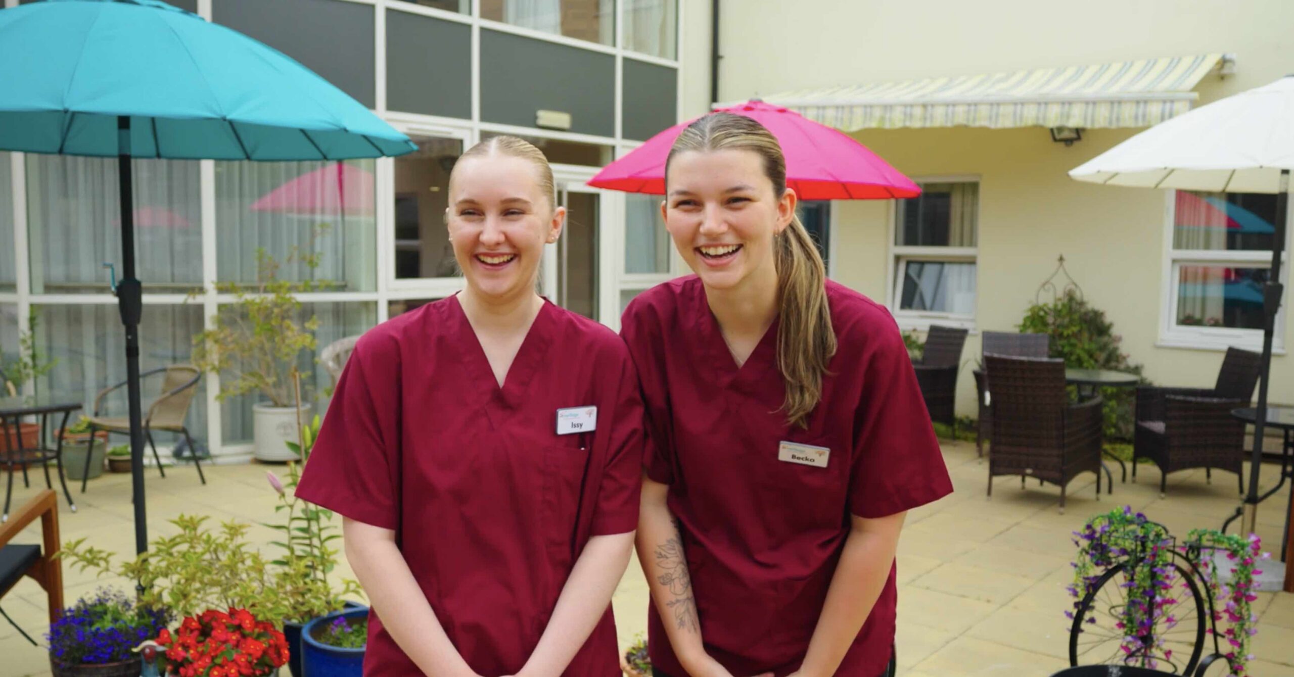Two female care workers in burgundy scrubs smiling off camera