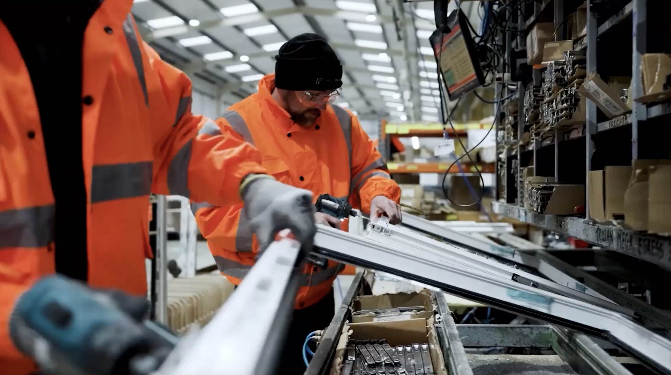 Two workers inside manufacturing facility working on window frames wearing orange hi-vis.